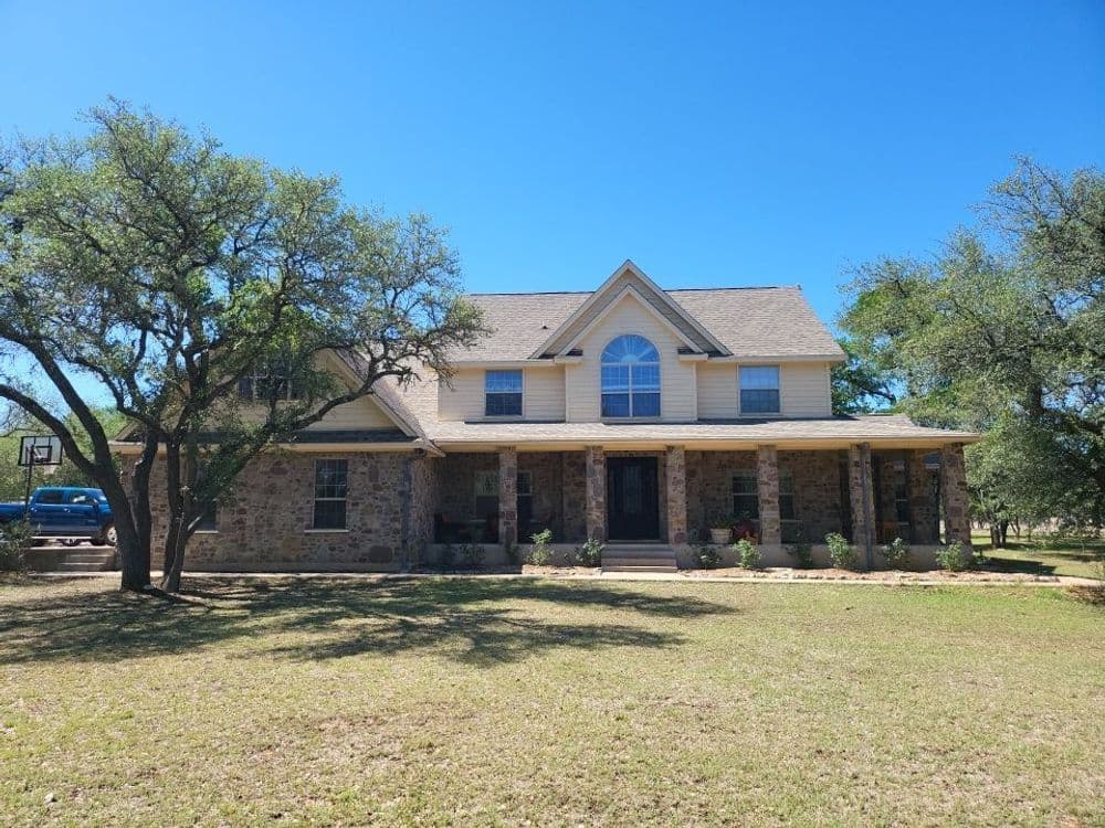Two-story stone house with a porch, surrounded by trees on a clear blue sky day.