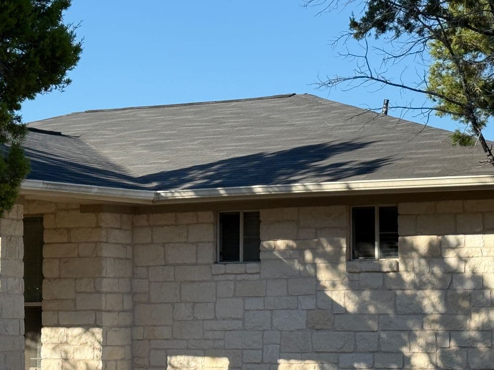 Gray shingled roof on a stone house with clear blue sky and tree shadows.