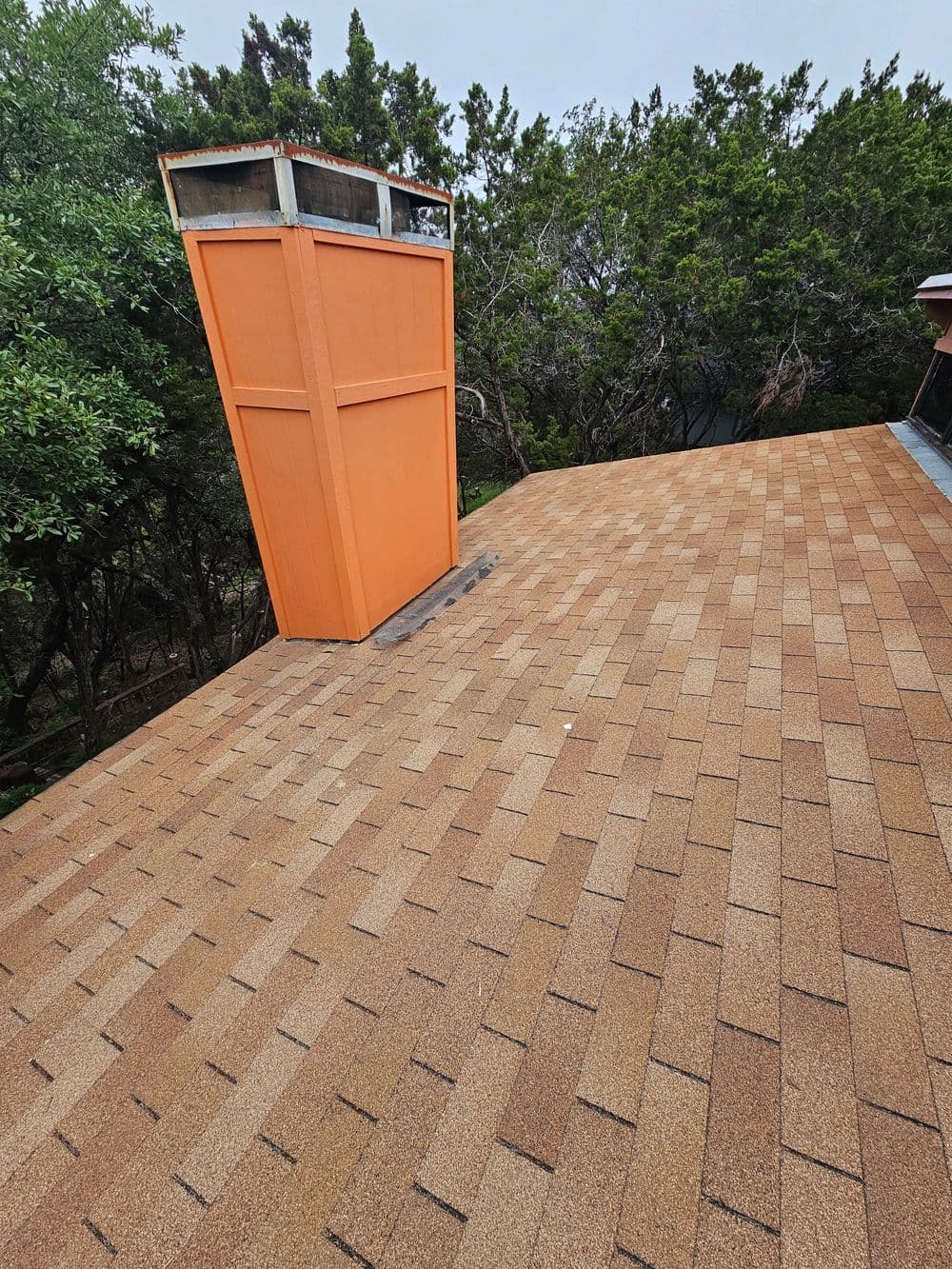 Roof with asphalt shingles and a distinctive orange chimney in a wooded area.