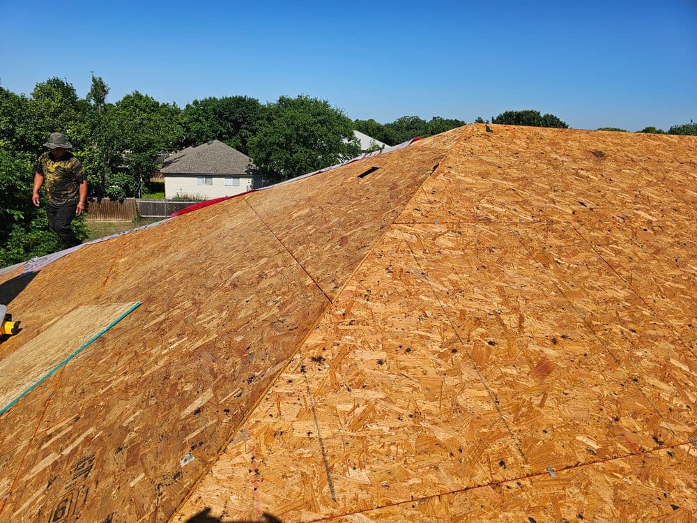 Construction worker installing OSB sheathing on a steep roof under blue sky.